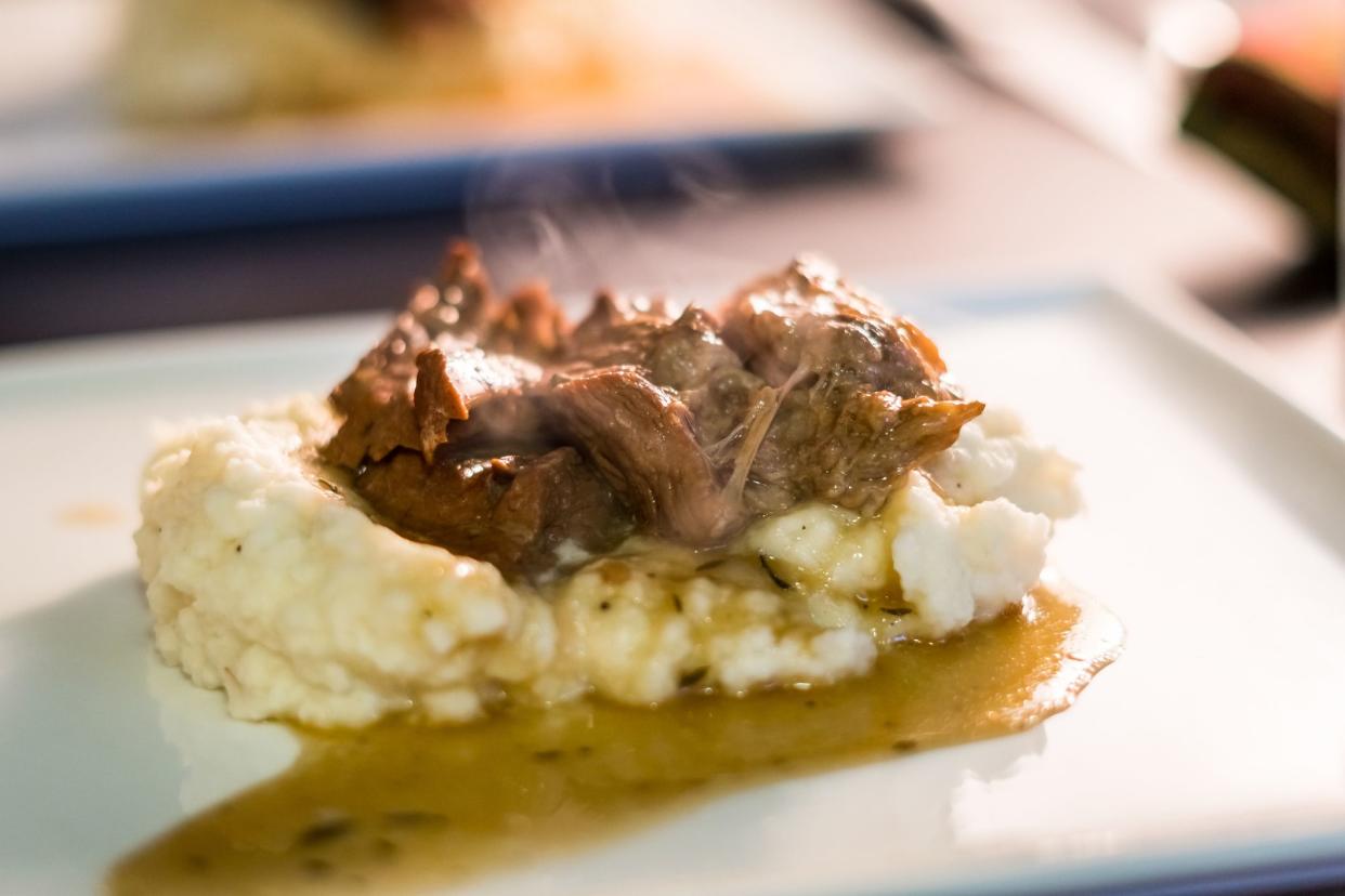 Juicy pot roast on top of whipped mashed potatoes with steam rising off the freshly cooked pot roast and Au Jus. Served on a square white plate.