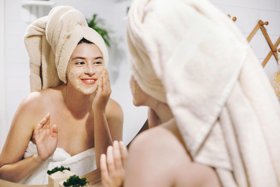 Skin Care concept. Young happy woman in towel making facial massage with organic face scrub and looking at mirror in stylish bathroom. Girl applying scrub cream, peeling and cleaning skin