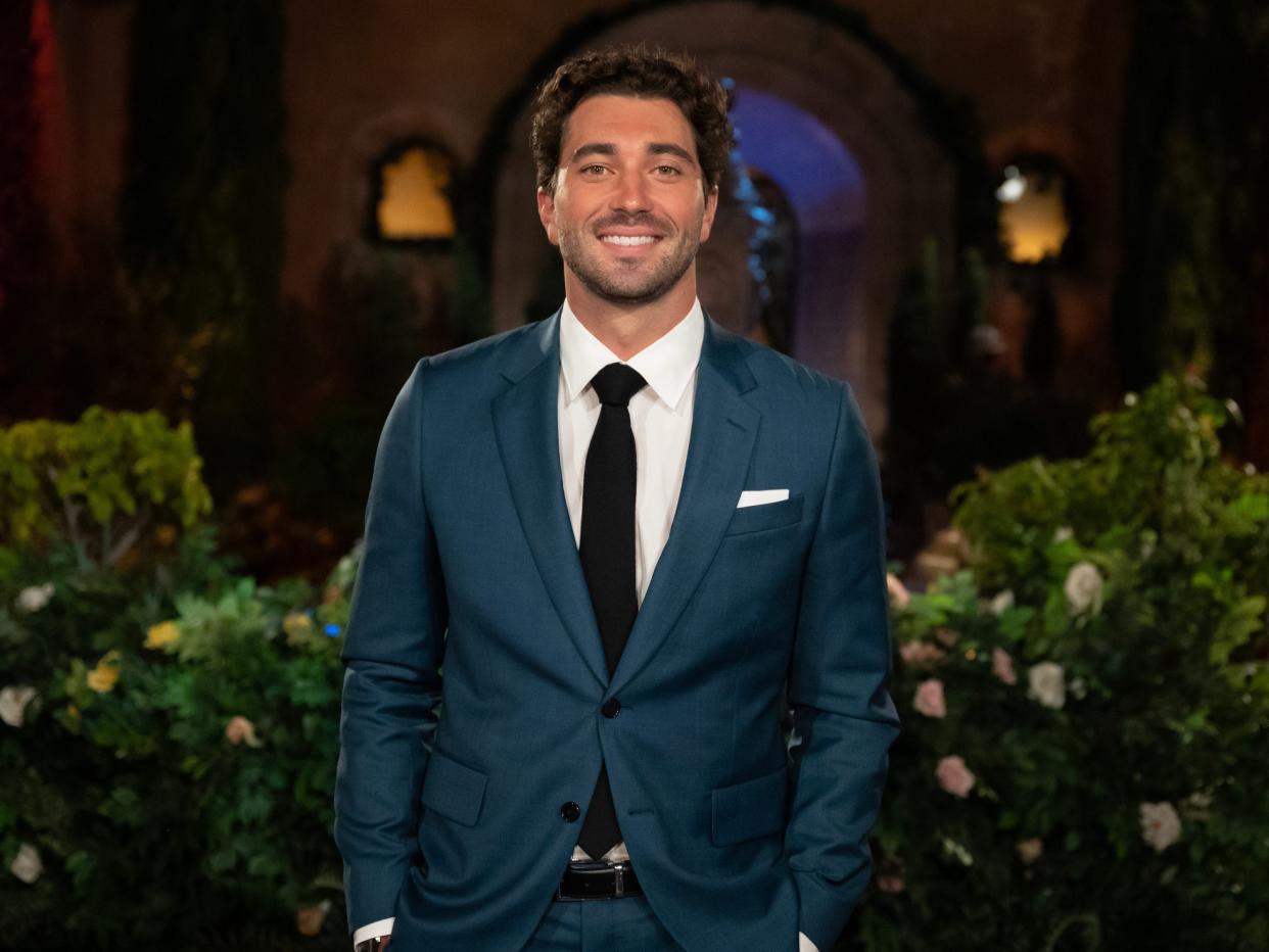 Joey Graziadei smiles in front of the Bachelor mansion in a blue suit, white shirt, and black tie.