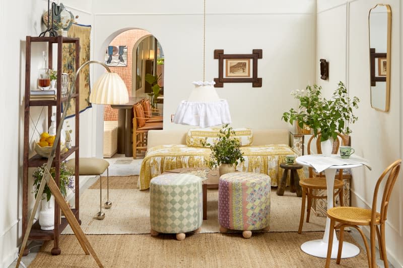 Head on view of a living room with white walls, a rattan rug.  Against the back wall is a low, beige couch with yellow and white ruffled cushion, in front of the couch is a low wooden coffee table, and 2 patch work stools.