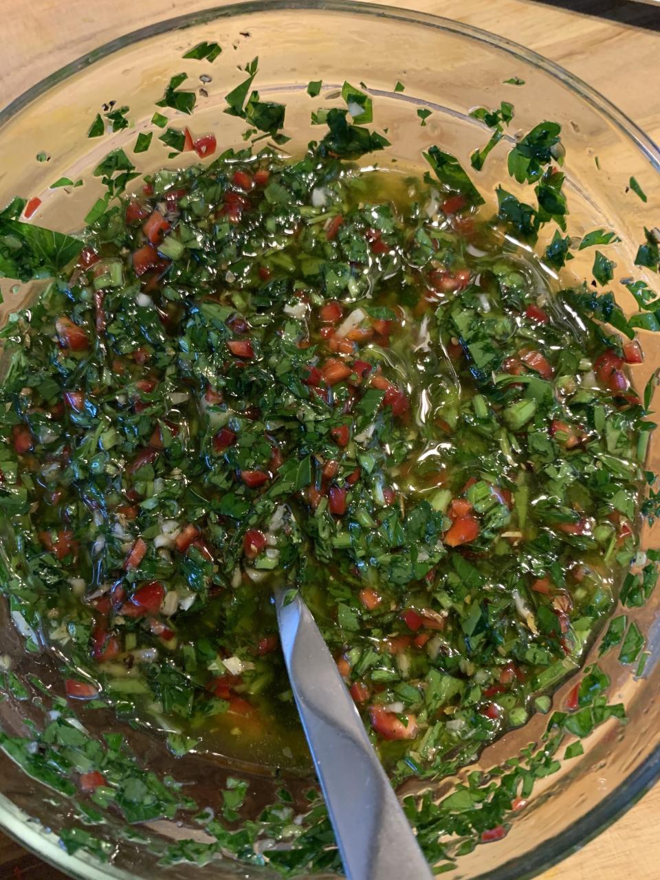 A clear bowl filled with freshly chopped herbs, garlic, and red pepper mixed with oil. A spoon is placed in the bowl, ready for serving