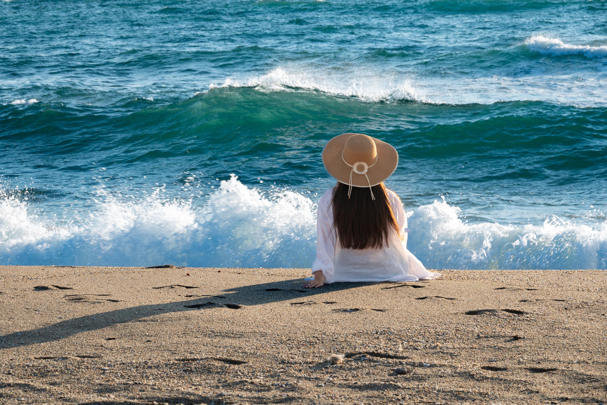 Rear view of an unrecognizable young woman with summer hat hat, sitting on the beach and watching waves and stormy sea. Nostalgic scene.