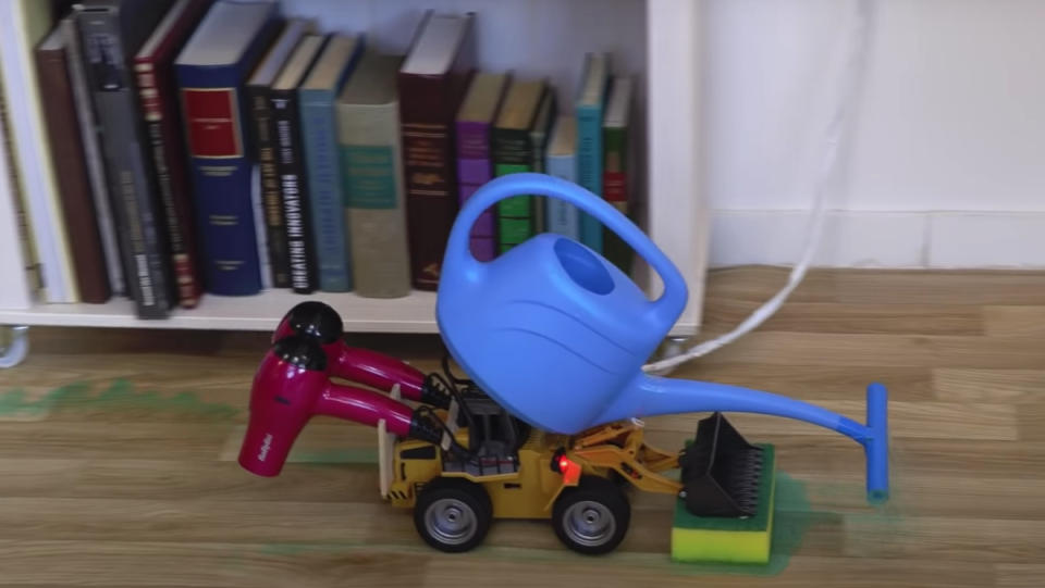 A Rube Goldberg cleaning contraption consisting of an RC car, watering pot and hair dryers cleaning up a mess on a wood floor.