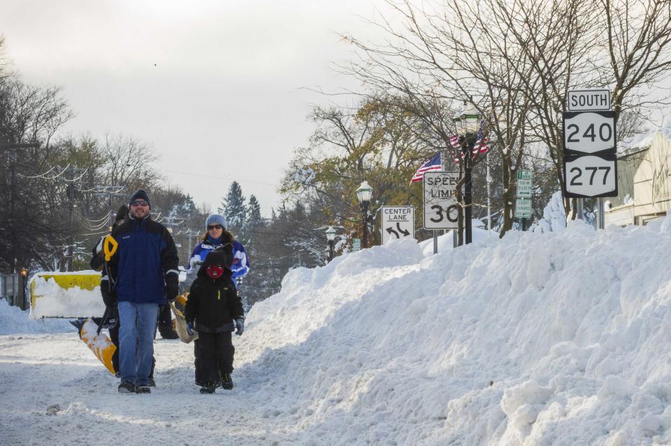 People carrying shovels walk in the street beside tall snowbanks in Buffalo