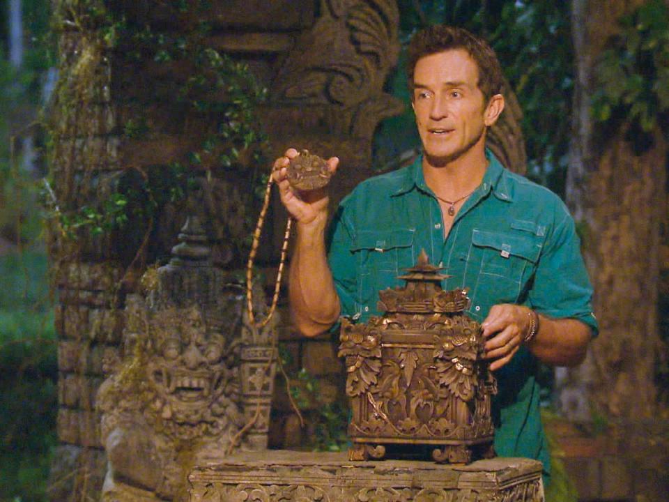 Jeff Probst in a blue shirt holding up a hidden immunity idol on Survivor