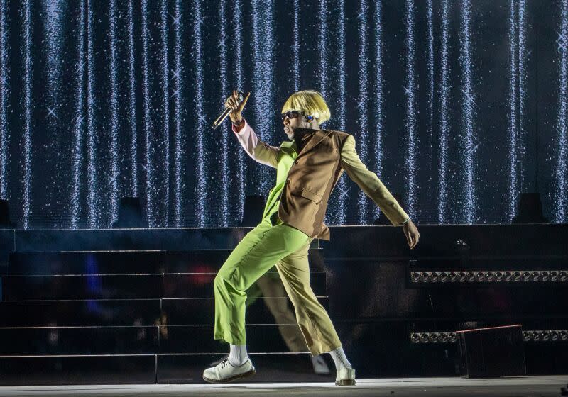 Tyler, the Creator in a platinum blonde bowl cut wig and a green an brown suit posing on a stage