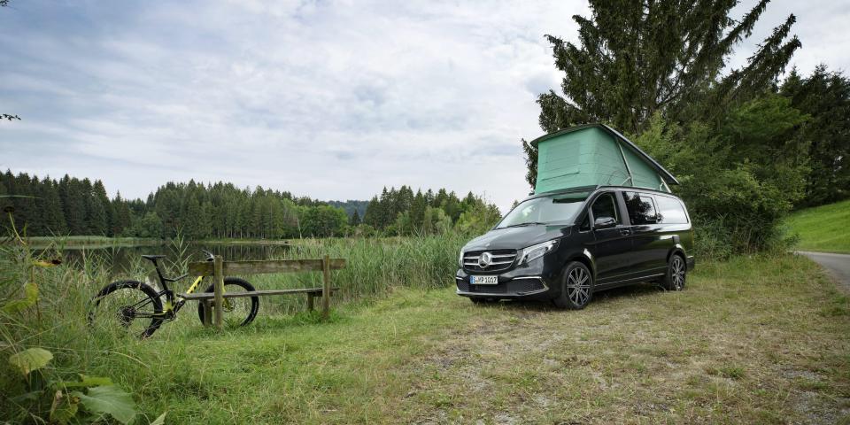 Mercedes-Benz's Marco Polo camper van RV with a pop-top roof sitting on a grassy patch in front of a tree. There's a bench with a bicycle near the van.