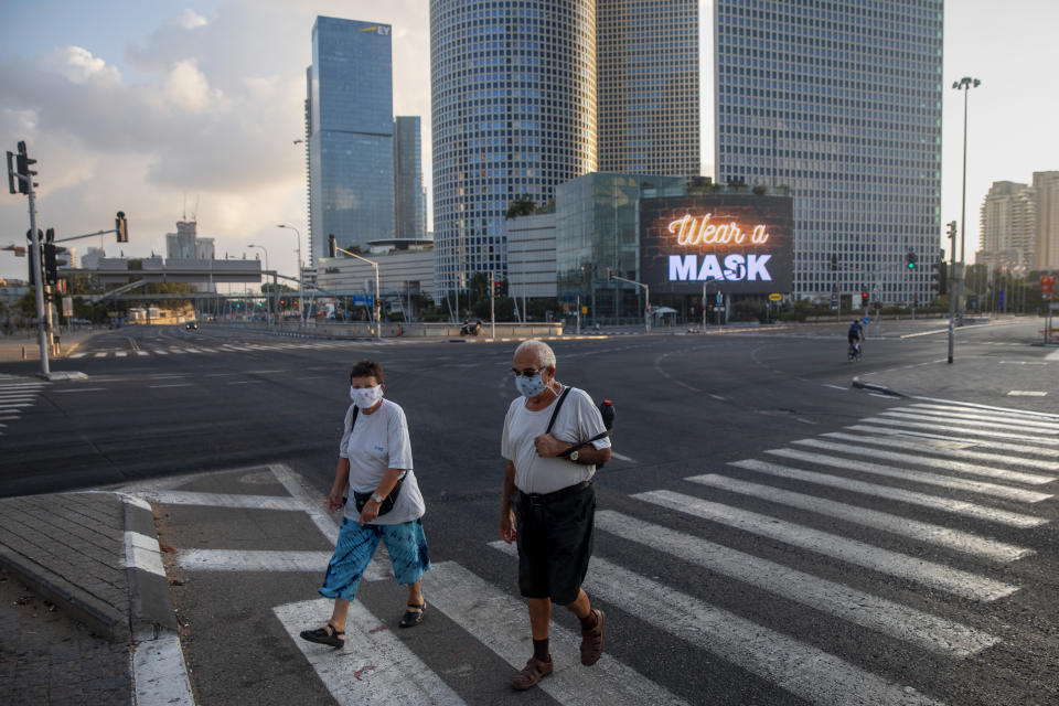 People wearing face masks cross a road next to a billboard calling people to wear masks on a mostly empty street following new restrictions in the three-week nationwide lockdown, in Tel Aviv, Israel, Saturday, Sept. 26, 2020. (AP Photo/Oded Balilty)