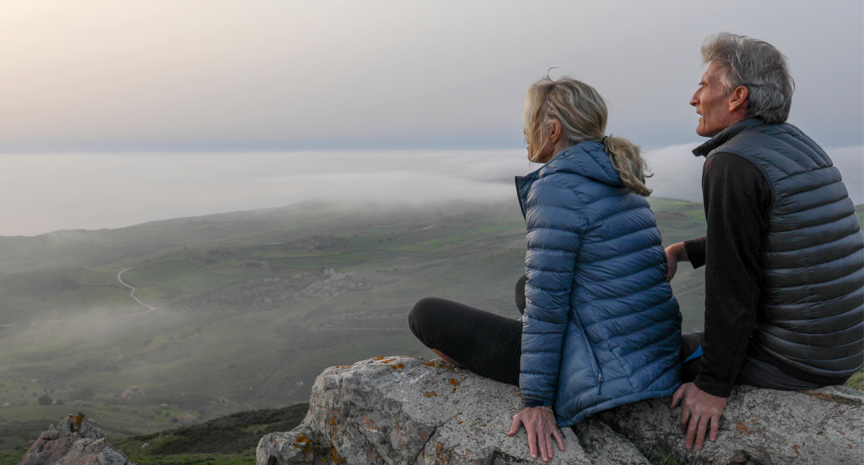 two people wearing jackets, man with grey hair and woman sitting on mountain rock looking at view wearing puffer jackets, amazon canada battery-powered heated jacket