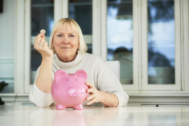 portrait of happy caucasian senior woman saving euro coin into piggybank and smiling at camera