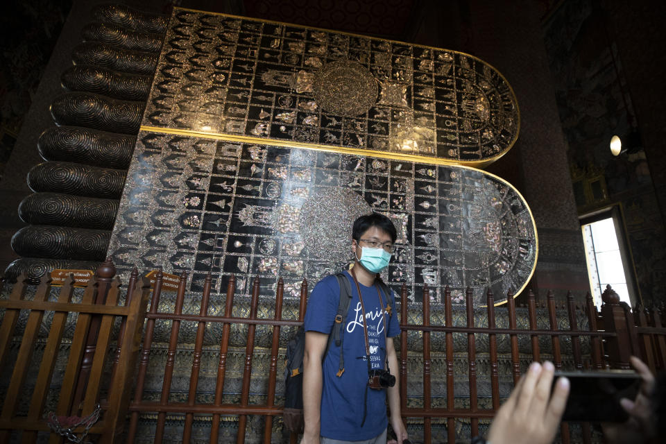 In this Feb. 7, 2020, photo, a masked tourist poses for a souvenir photo at Wat Pho in Bangkok, Thailand. There were less crowds in Bangkok&#39;s popular Wat Pho, a centuries-old Buddhist temple known for its giant reclining buddha, due to the virus scare. The complex of temples is normally visited by thousands of tourists. (AP Photo/Sakchai Lalit)