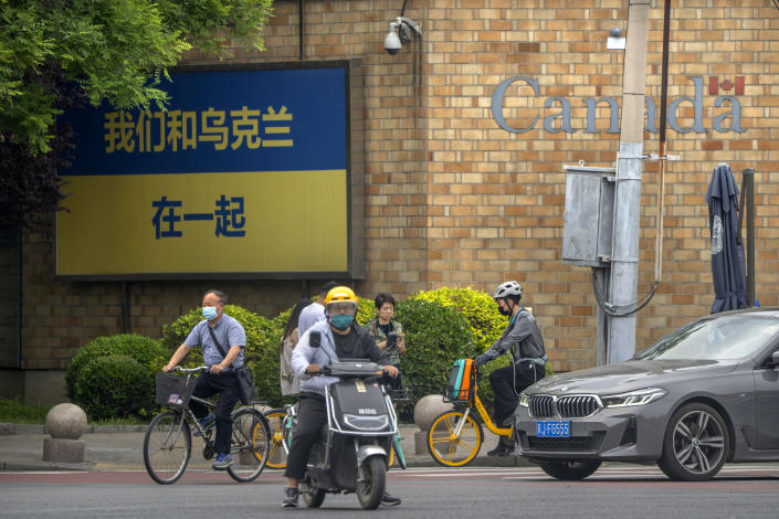 People riding bicycles and scooters wait to cross an intersection near a sign reading "We stand with Ukraine" outside the Canadian Embassy in Beijing, Wednesday, May 17, 2023. Embassies in Beijing have been asked by the Chinese government to avoid displaying propaganda after some raised Ukrainian flags or set up placards declaring support for Ukraine. (AP Photo/Mark Schiefelbein)