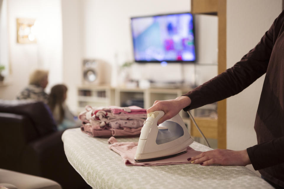 Someone irons clothes on an ironing board while a child and elderly person sit on a couch in the background watching TV