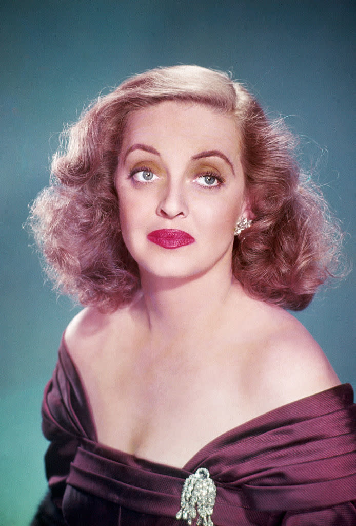 Bette Davis poses in an off-the-shoulder gown with a brooch, styled with elegant waves in her hair and bold makeup