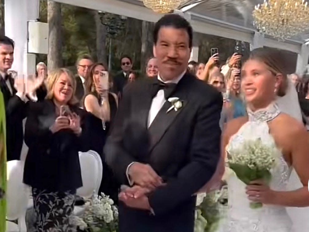 Lionel Richie, in a black tuxedo, smiles as he walks daughter Sophia Richie, in a white high-necked dress wearing a veil and carrying a bouquet, down the aisle at Sophia's wedding, while wedding guests applaud in the background.