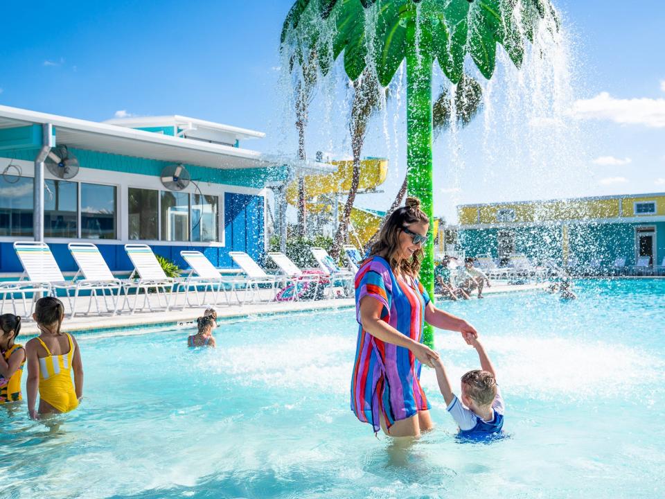 A woman and child playing in a shallow pool with other people. There's a fake tree spraying water behind them.