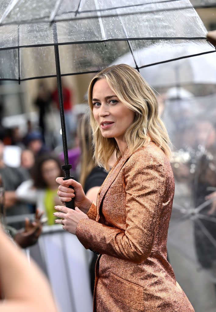 emily blunt looks at the camera while holding the handle of a large clear umbrella, she wears a golden suit jacket