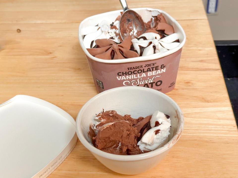 A brown tub of chocolate and vanilla gelato swirled with fudge sitting next to a white bowl of gelato on a wooden table