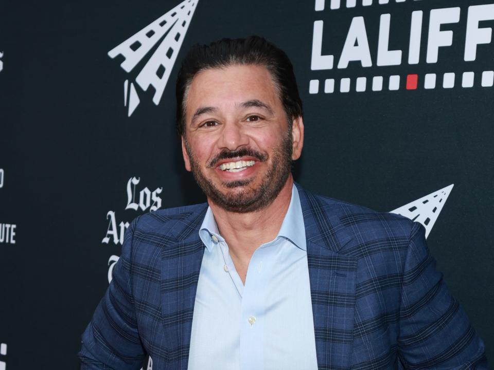 al madrigal, a middle aged man with dark brown hair and trimmed brown facial hair. he's smiling and holding his hands on his hips, wearing a blue checkered blazer and light blue shirt.