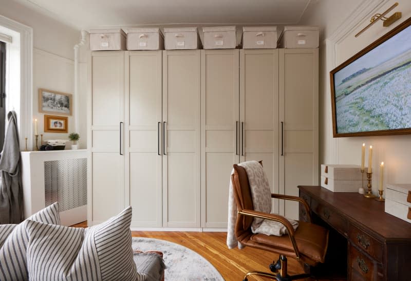 Wall of closets topped with storage boxes in bedroom with framed television above wood desk.