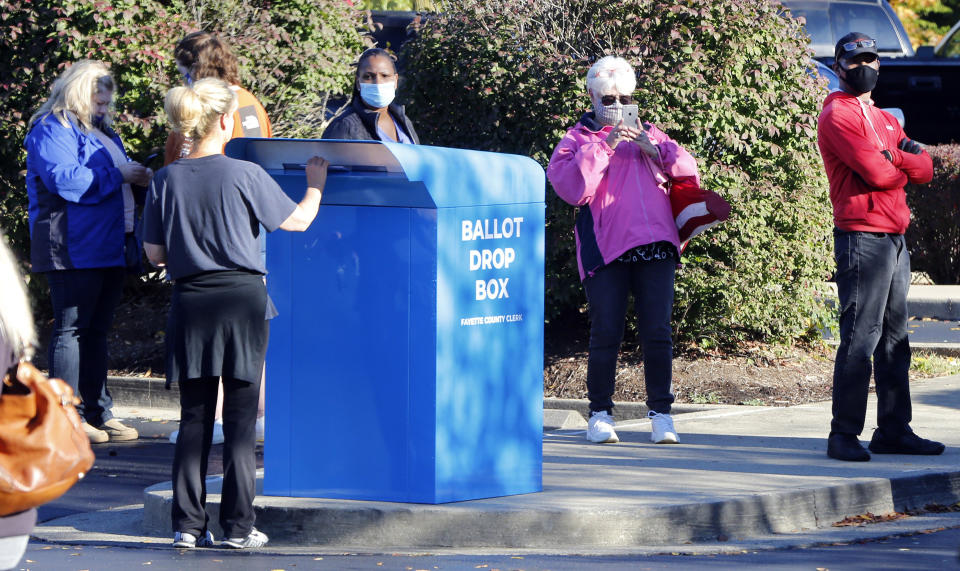 Liz Young drops off her ballot at the Tates Creek library while voters wait in line to vote in-person Tuesday, Oct. 13, 2020 on the first day of early voting for the 2020 election in Fayette County. Tens of millions of Americans already cast ballots in the 2020 election amid record-breaking early voting during the coronavirus pandemic. In Kentucky the Democratic governor and Republican secretary of state struck an agreement to make it safer to vote. For the general election, the state is offering absentee voting to anyone who feels at risk from COVID-19, as well as early in-person voting to try to hold down crowds on Election Day. (Brian Simms/Lexington Herald-Leader via AP)