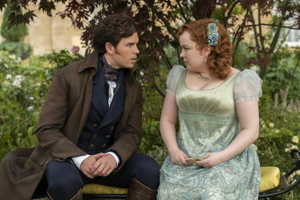 Luke Newton and Nicola Coughlan in period costumes, sitting on a bench, looking at each other intently, in a garden setting