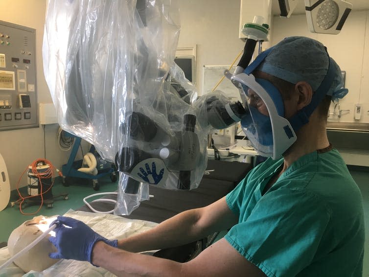 A surgeon in scrubs and a hairnet wears a snorkel mask and looks down a microscope while simulating an operation on a skull.