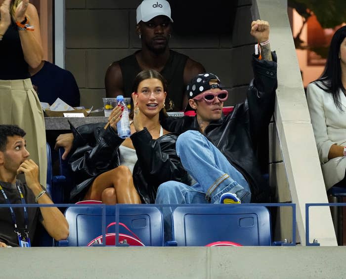 Justin Bieber lifts his arm to cheer the match as he sits next to his wife, Hailey; NBA player Jimmy Butler sits behind them