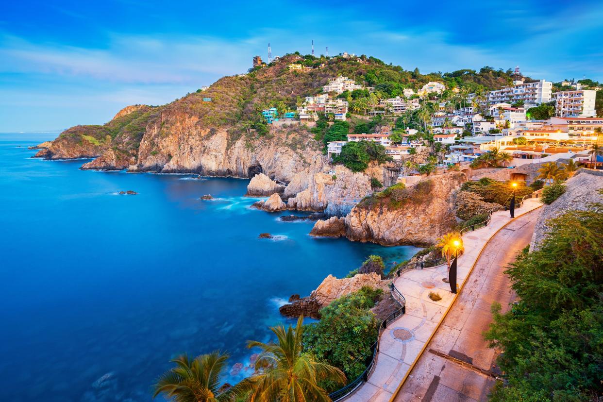 Rocky coastline with promenade in Acapulco, Mexico, several houses along the cliffs, during early evening with lights, against an expansive blue sky