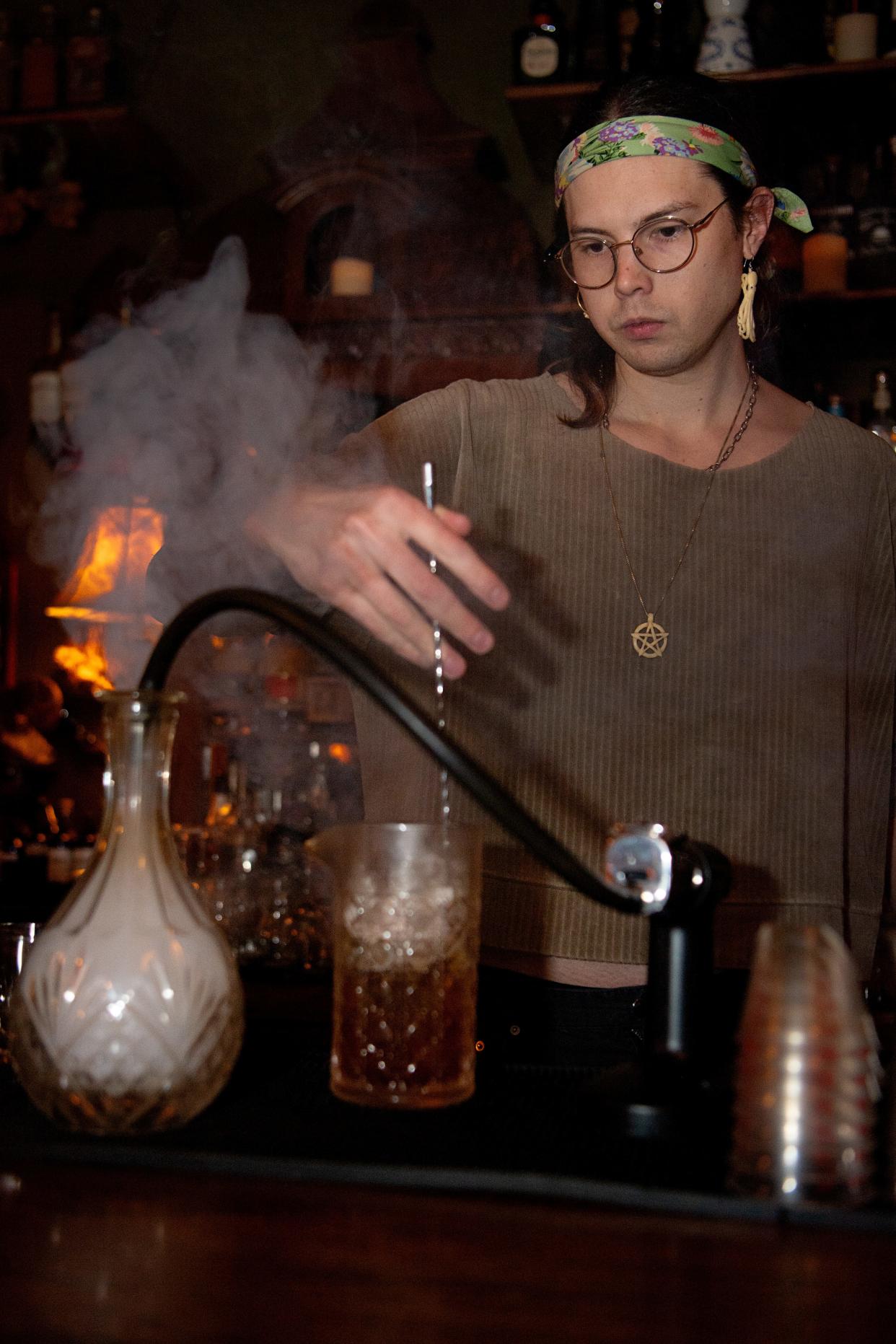 August Henson stirs ingredients for The Crow and Quill’s Fall Fashion, the cocktail bar’s take on an Old Fashioned, while preparing pipe tobacco smoke for the drink, April 4, 2024, in Asheville.