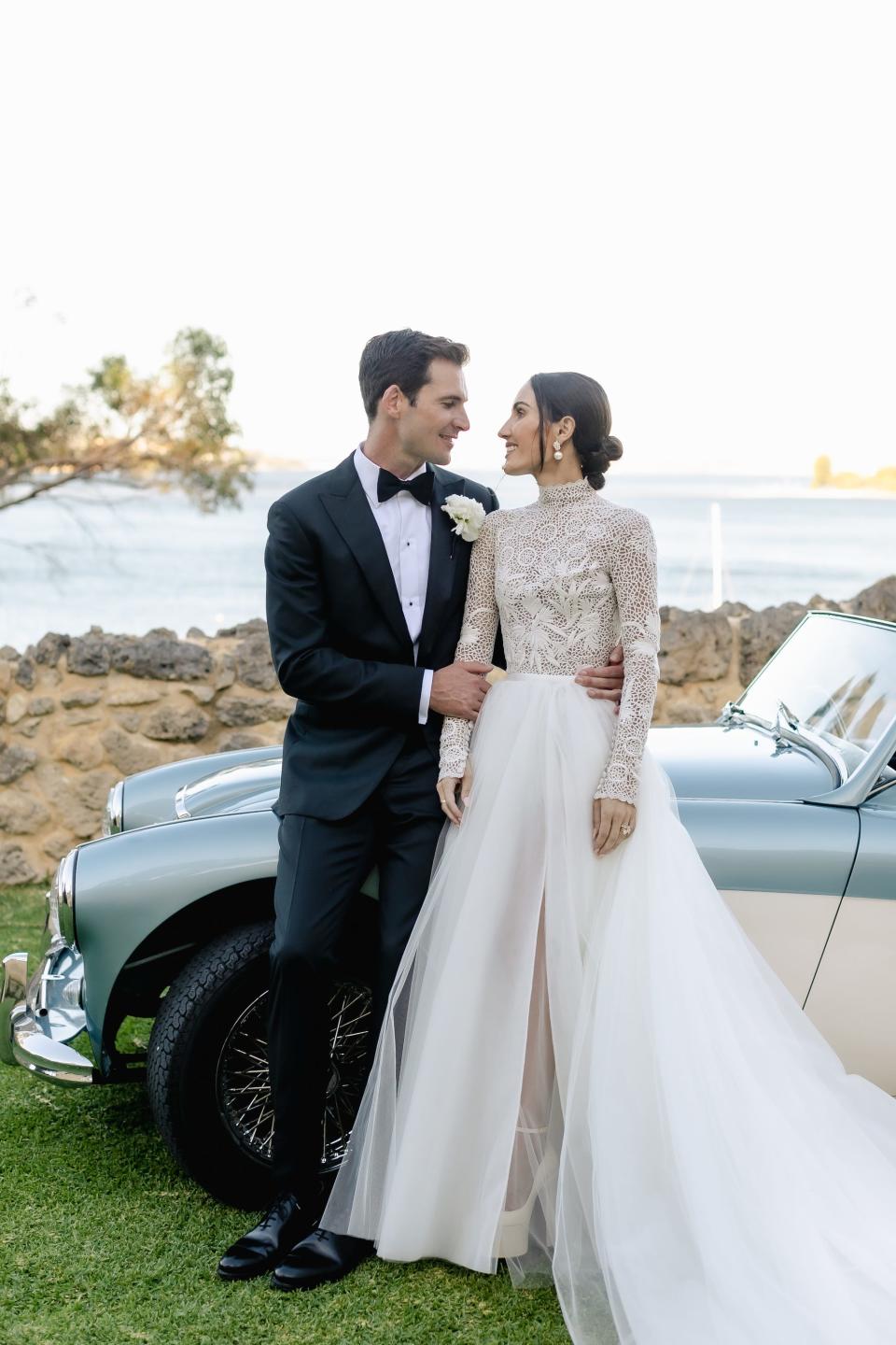 A bride and groom lean against a convertible and smile at each other in their wedding attire.