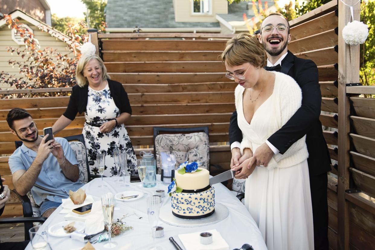 Millennial newlywed couple cutting the cake at wedding cocktail party in backyard. This is part of a series of a real wedding during Covid-19. Horizontal outdoors waist up shot with copy space.