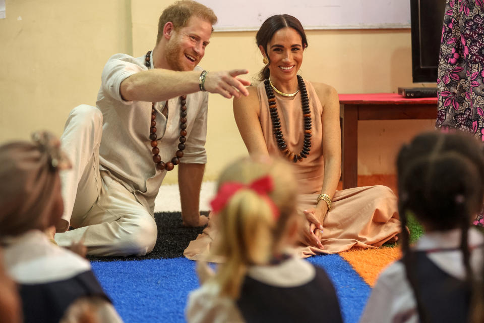 Prince Harry and Meghan Markle sitting, smiling at children at the Lighthouse Academy