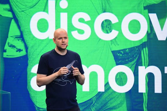 Spotify CEO Daniel Ek stands on stage at a press conference with a blue and green backdrop of people dancing