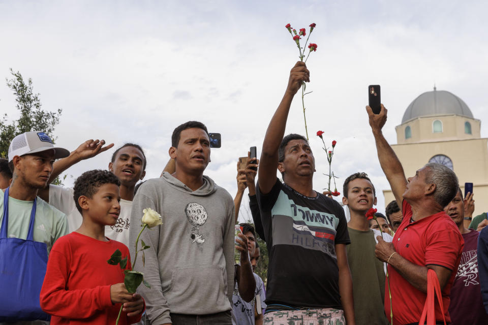 Migrants hold flowers and record speakers during a vigil for the eight migrants that were killed and several others that were injured the day before while waiting at a bus stop, in Brownsville, Texas, Monday, May 8, 2023. (AP Photo/Michael Gonzalez)