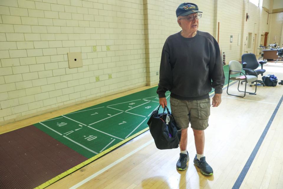 A man in a Korean War veteran's cap holds a gym bag near a shuffleboard.