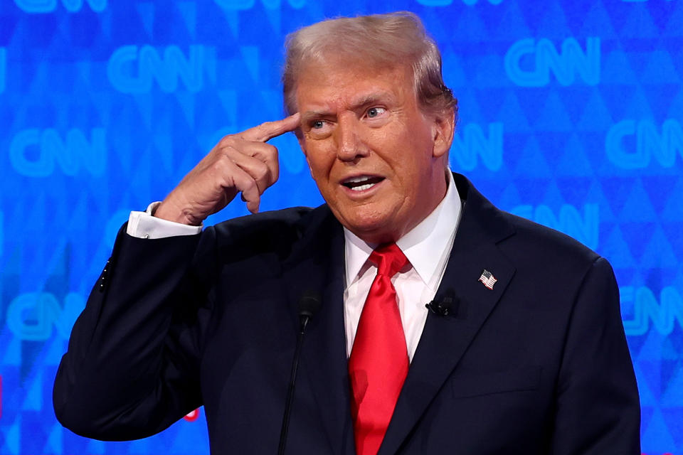 Donald Trump gestures during a CNN interview in front of a blue background. He wears a dark suit, white shirt, and red tie