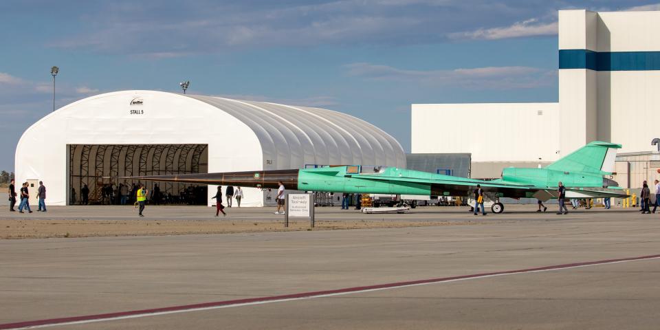 A picture shows NASA's X-59, a long green-colored jet with a very long black nose, near a hangar.