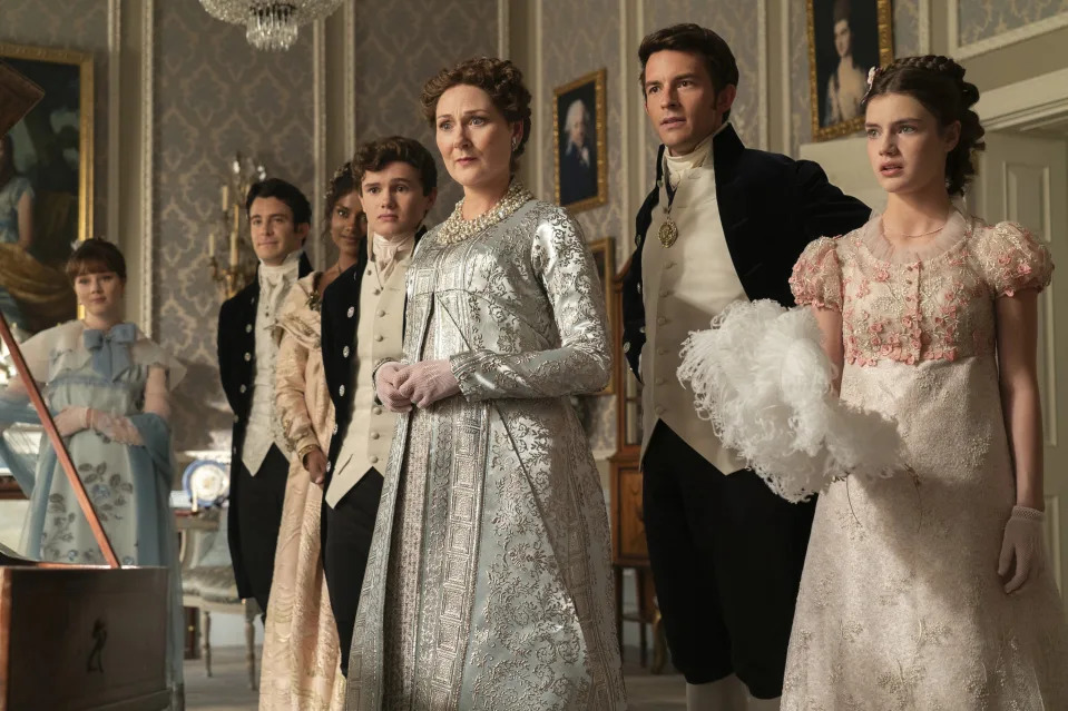 Ruth Gemmell surrounded by half a dozen others wearing period dress in an ornate room.