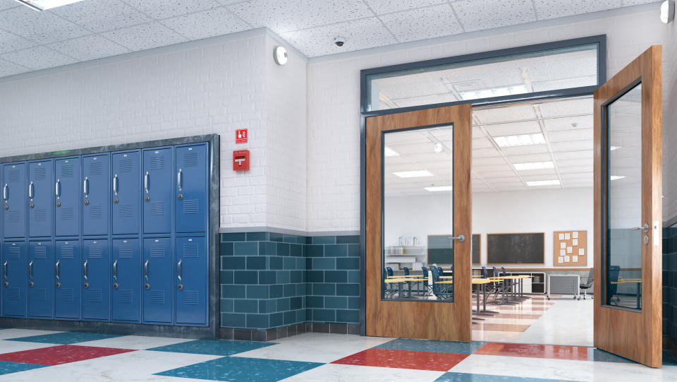 An empty school hallway with blue lockers and an open classroom door revealing desks and chairs inside the room