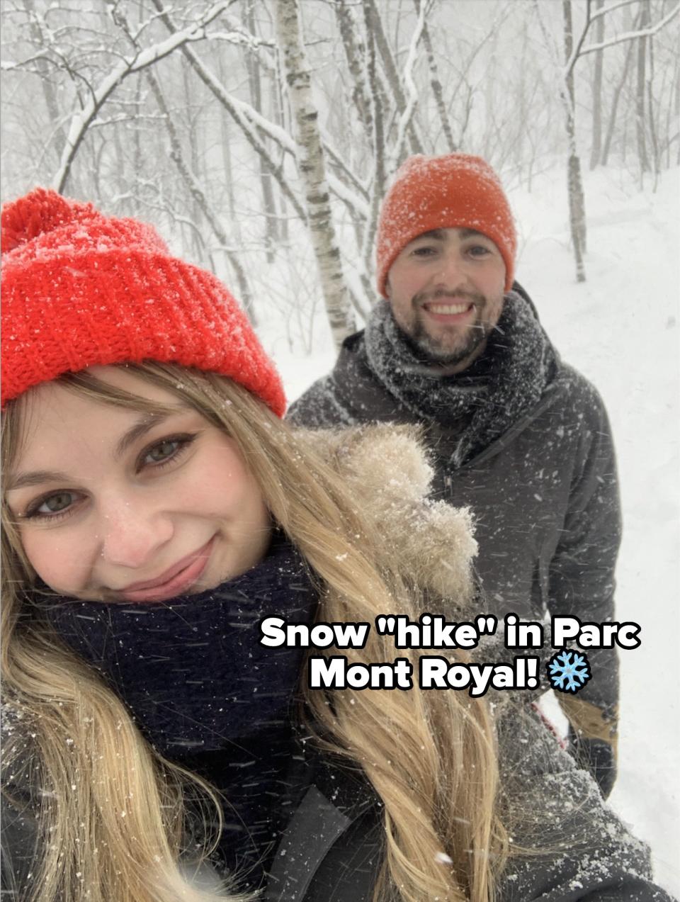 Man and woman smile for a selfie in a snowy forest, each wearing red hats. Trees and snow visible in the background