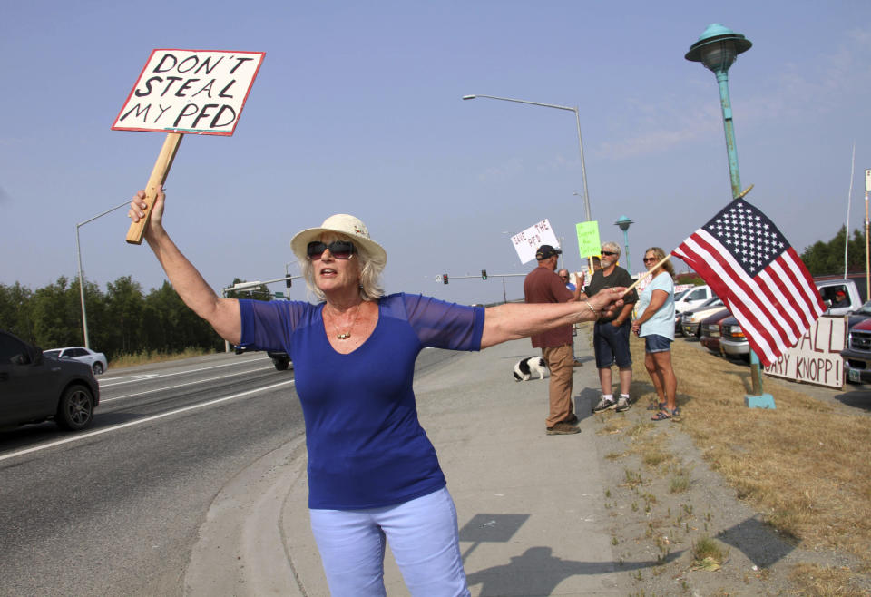 Katherine Hayes waves a flag and a sign urging Alaska lawmakers to fund a full oil wealth fund check, known locally as the PFD or Permanent Fund Dividend, Monday, July 8, 2019, in Wasilla, Alaska. Some Alaska lawmakers are meeting in Wasilla July 8 instead of Juneau, where state House and Senate leadership have decided to hold the special session, called to determine the amount of this year's check. (AP Photo/Mark Thiessen)