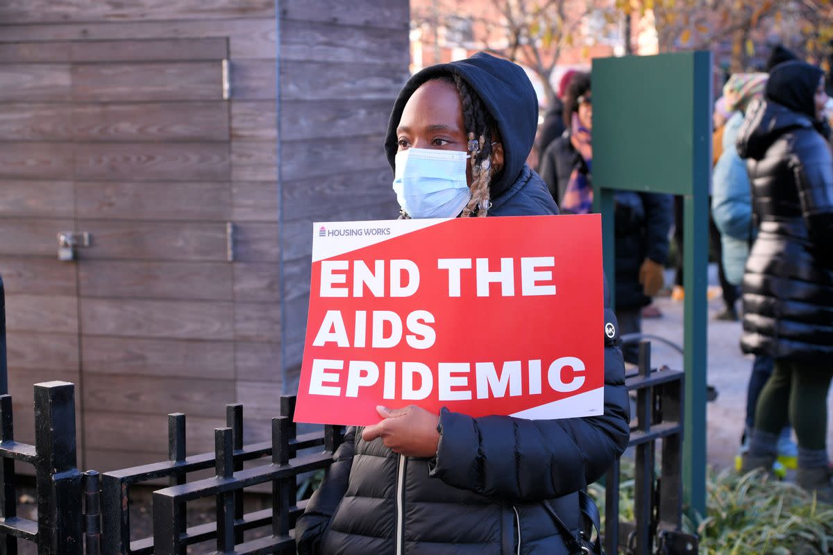 A Black person holds a red sign with white letters that say "END THE AIDS EPIDEMIC.". 