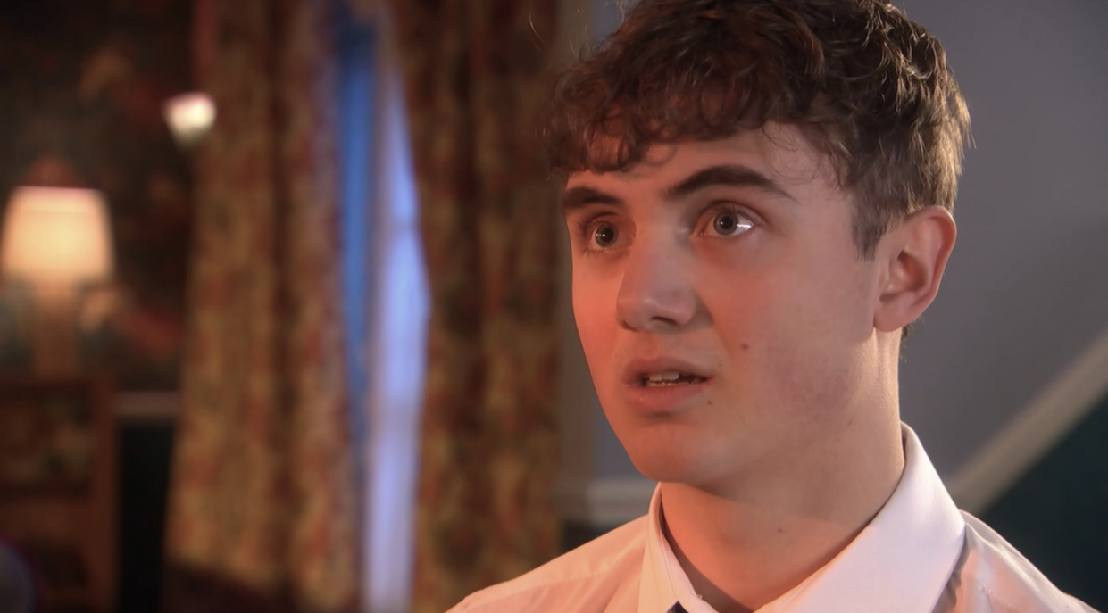 jj osbourne in hollyoaks, a teenage boy stands looking at someone, he has short curly brown hair and is wearing a school uniform