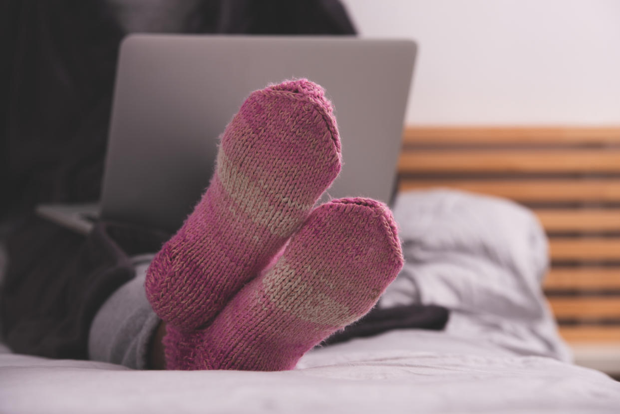 Close up image of pink, purple cozy soft warm knitted winter socks on woman's legs. The unrecognizable woman lying in bed, using a laptop.