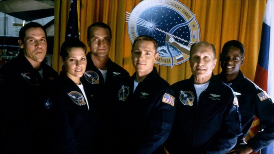 From left to right, the cast of Deep Impact (1998): Jon Favreau, Mary McCormack, Aleksandr Baluev, Ron Eldard, Robert Duvall, and Blair Underwood. All are wearing a dark blue space jumpsuit and standing in front of an orange curtain and a space emblem.