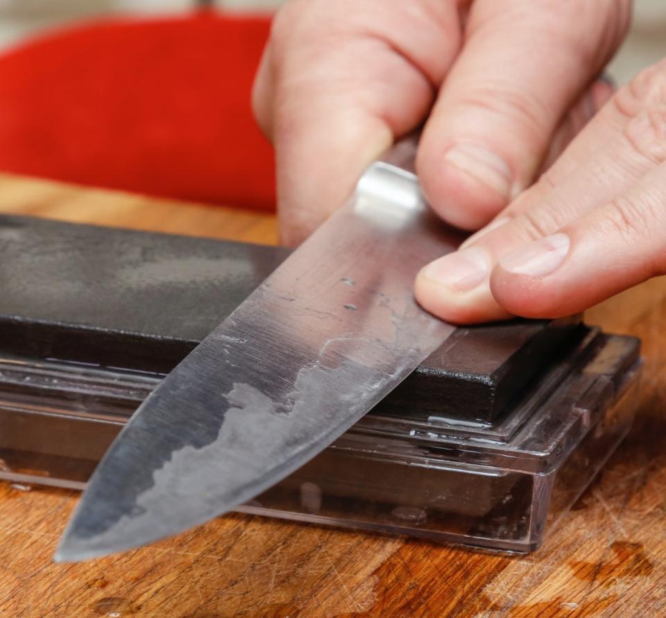 Close-up of hands sharpening a kitchen knife on a whetstone placed on a wooden cutting board