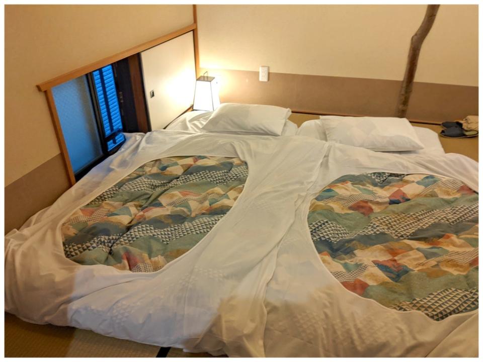 A Japanese-style futon bed on a tatami mat floor next to a small floor-level window. The bed has a colorful duvet cover on top.