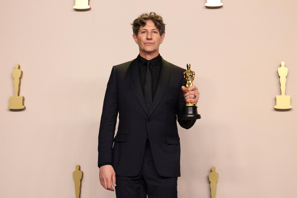 Person holding an Oscar trophy, dressed in a black suit and bowtie, standing in front of statue silhouettes