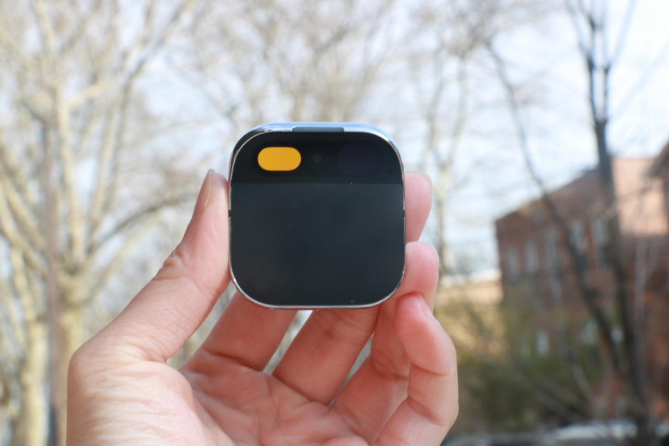 The Humane AI Pin held in mid-air in front of some bare trees and a street with red brick buildings on it.